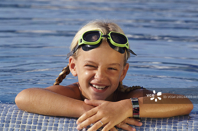 女孩的头和肩膀靠在泳池边，额头上戴着护目镜，微笑着图片素材