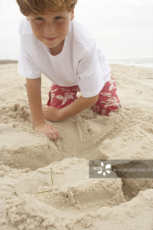 男孩(6-8)跪在沙滩上玩沙子，面带微笑，画像图片素材