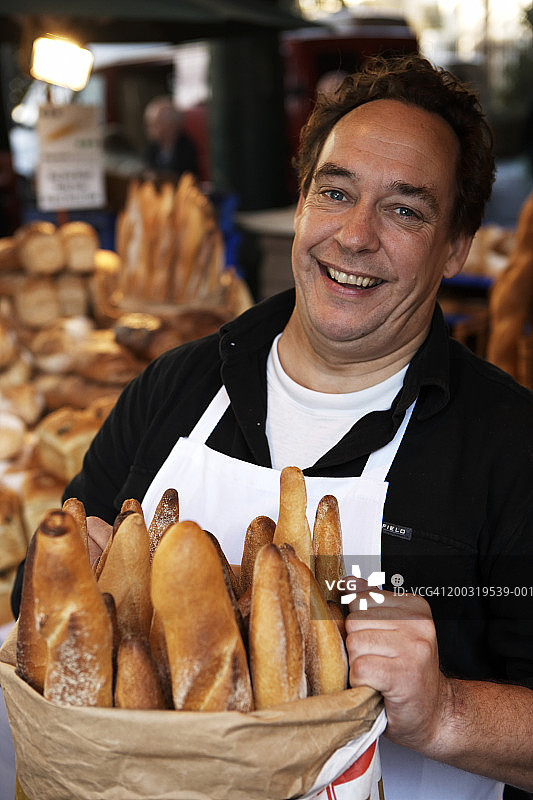 市场摊贩拿着一袋法棍面包，面带微笑，画像图片素材