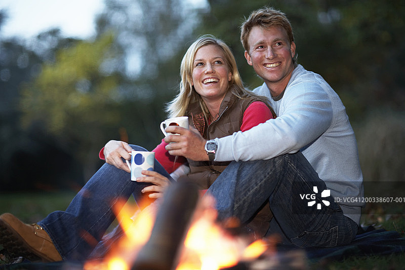 一对年轻夫妇坐在火边，拿着杯子，微笑着图片素材
