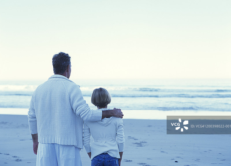 男子挽着女孩(10-12)站在海滩上，后视图图片素材