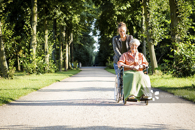 一个年轻女子推着一位坐轮椅的老妇人穿过公园图片素材