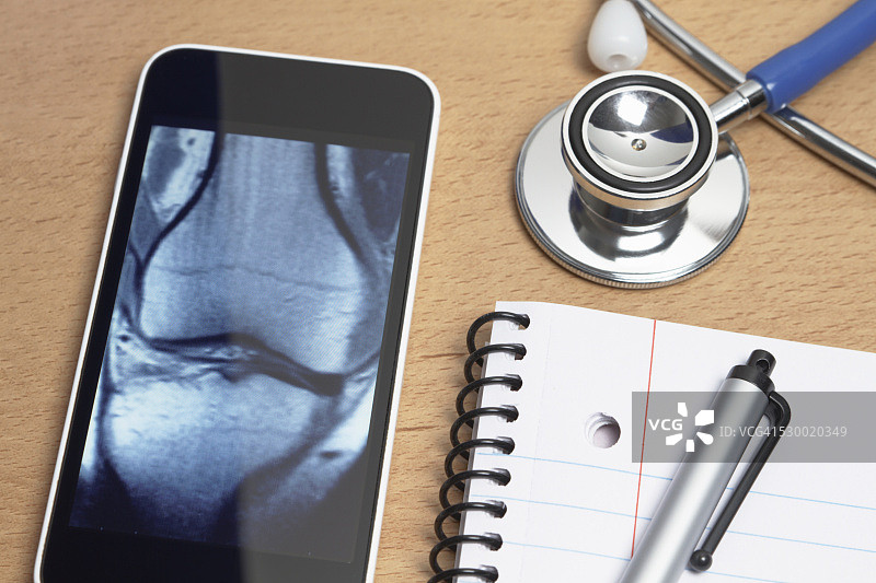 医疗保健中的技术使用。智能手机显示膝盖的x射线CT扫描、听诊器、记事本和医生办公桌上的笔图片素材