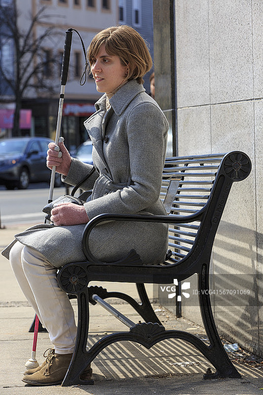 年轻的盲人妇女拄着拐杖坐在镇上的长椅上图片素材