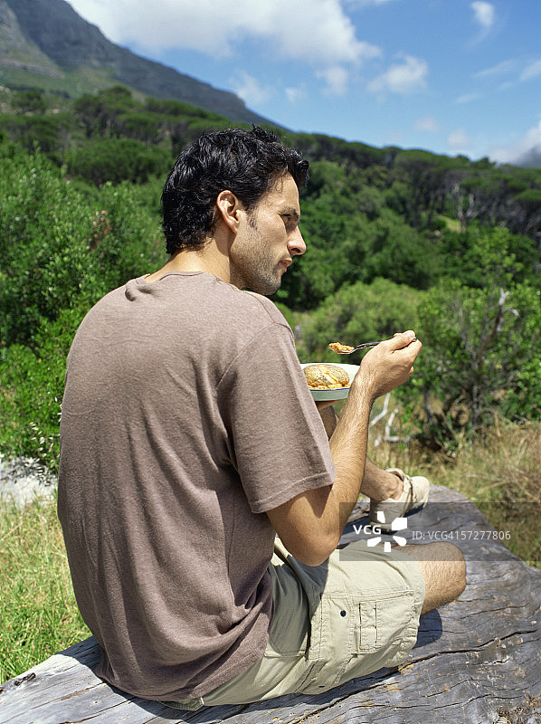 一个年轻人的后视图，坐在一块岩石上吃东西图片素材