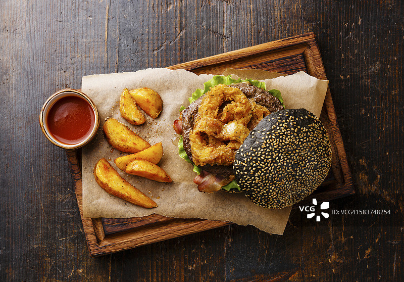 黑色的汉堡配以芝麻小圆面包、肉、培根、洋葱圈薯条和薯角放在深色的木质背景上图片素材