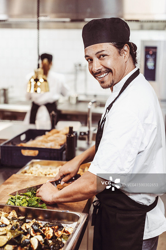 侧视图肖像快乐的男厨师切叶蔬菜在商业厨房图片素材