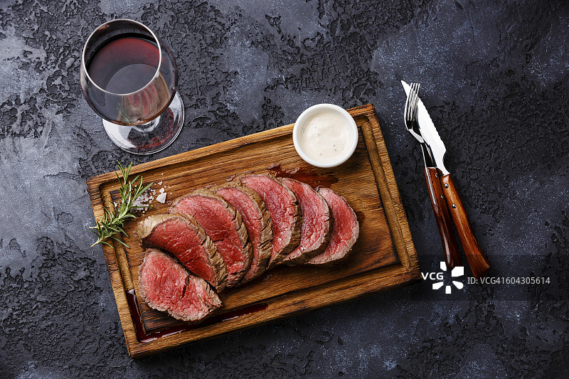 切成薄片的烤里脊牛排、烤牛肉和胡椒酱放在木砧板上，红酒在深色的背景上图片素材