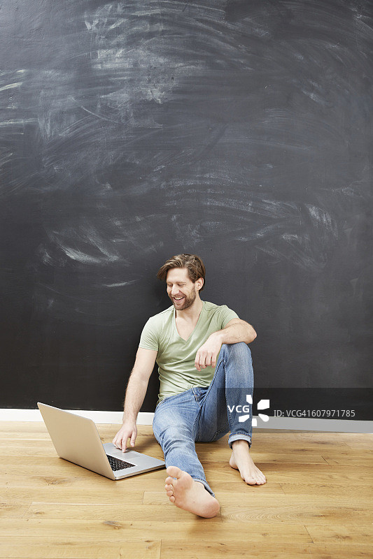 微笑的年轻人坐在黑板前用笔记本电脑的肖像图片素材