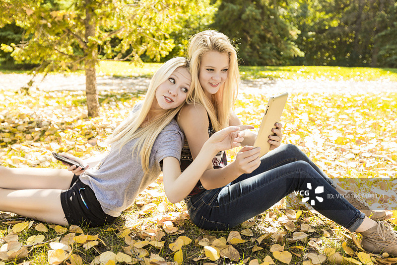 秋天，两姐妹在城市公园的户外玩，在他们的智能手机和平板电脑上浏览社交媒体图片素材
