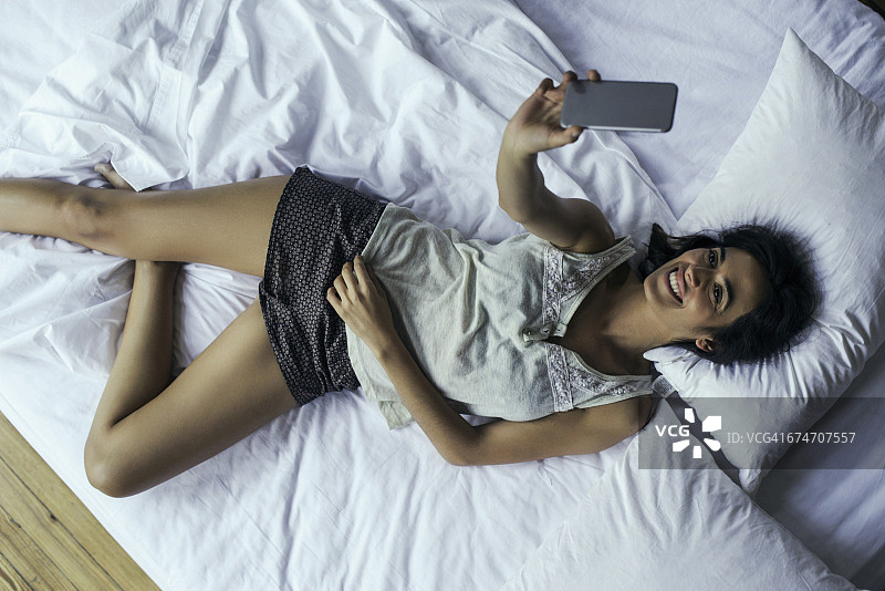 女人躺在床上用智能手机自拍图片素材