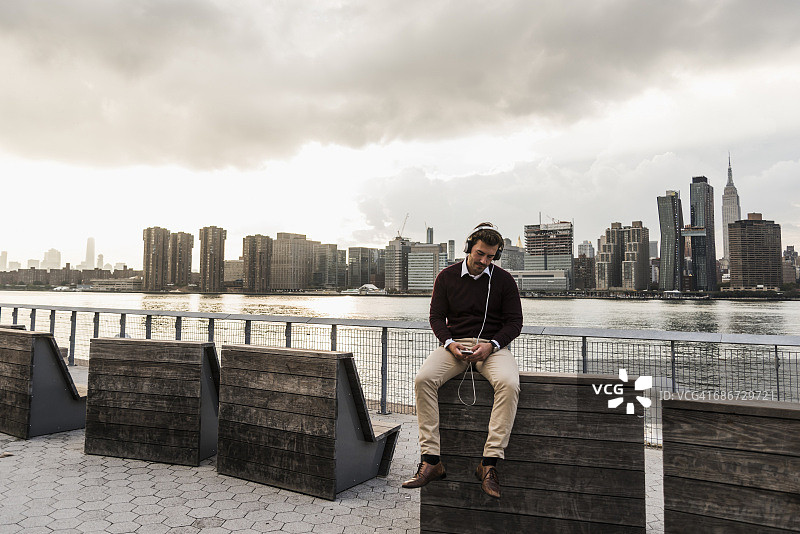 美国，纽约市，一个年轻人戴着耳机和手机坐在东河图片素材