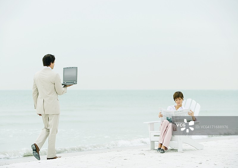 一个女人坐在沙滩上看报纸，商人拿着打开的笔记本电脑向她走来图片素材