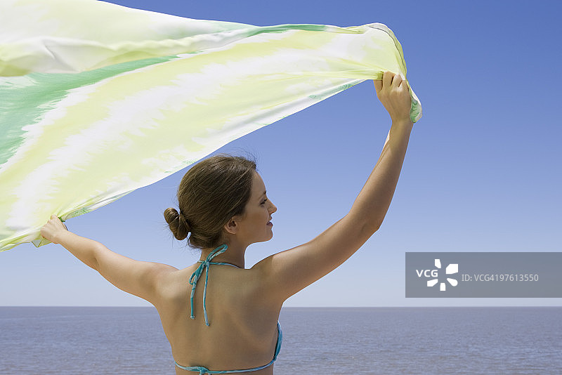 年轻女子站在大海前，在微风中拿着围巾图片素材