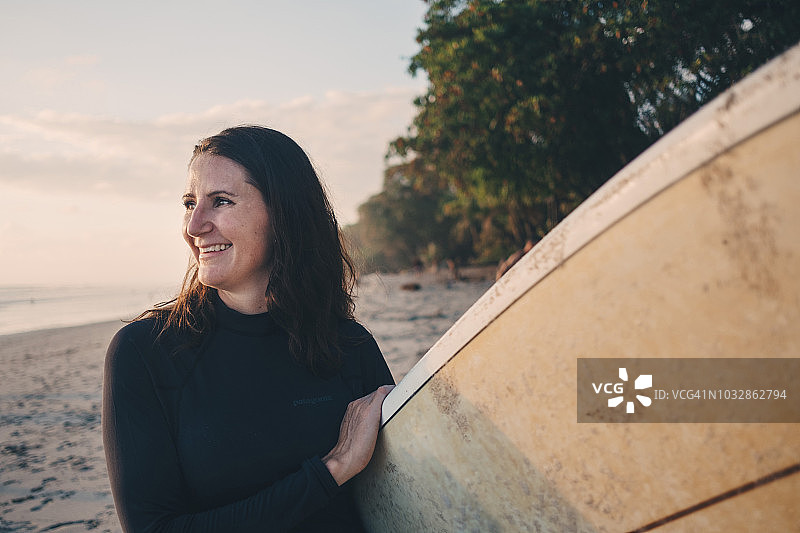 微笑的成熟女人背着冲浪板在海滩上图片素材