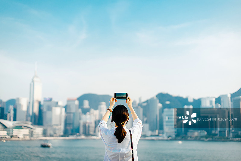 后视图的女人享受维多利亚港的壮观景色，并在一个阳光明媚的日子用智能手机拍摄香港城市天际线图片素材