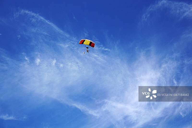 以白云和蓝天为背景的红黄相间的降落伞和跳伞者的剪影。图片素材