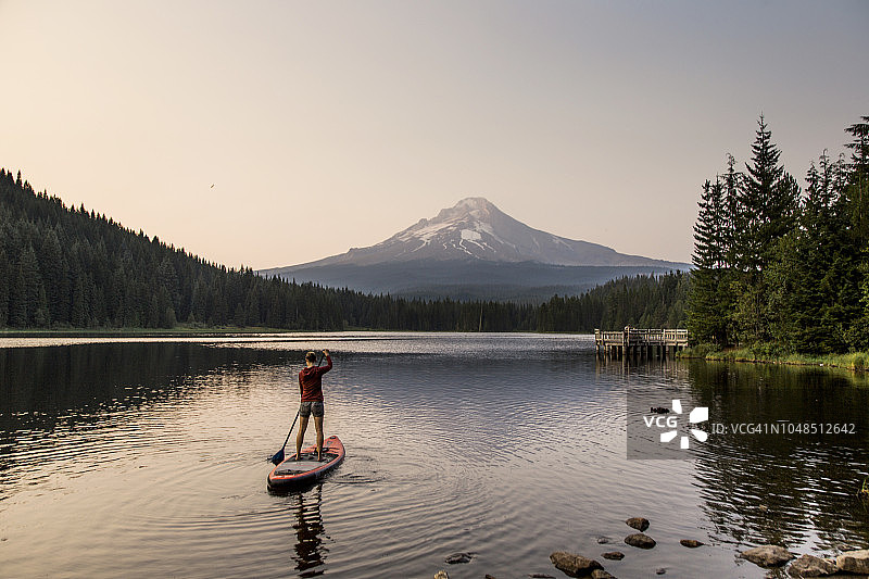 一个年轻的女人划着她的立式桨板(SUP)在一个高山湖泊在一个火山基地在日落在俄勒冈州。图片素材