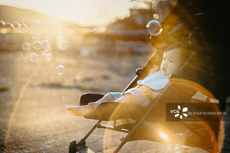 在美丽的日落时分，小女孩坐在婴儿车里玩着漂浮的泡泡图片素材