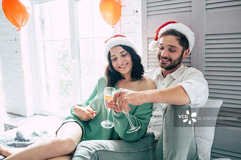 幸福的年轻夫妇戴着圣诞帽拥抱亲吻，在明亮的卧室里喝着香槟庆祝圣诞节图片素材