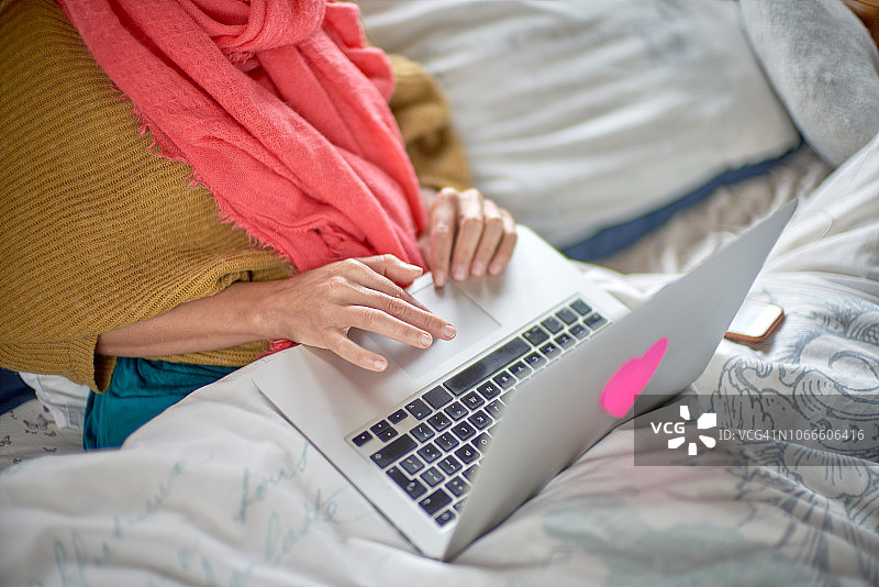 一个女人坐在家里的床上用她的笔记本电脑工作。图片素材