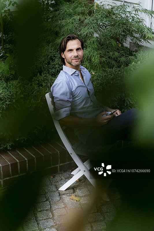 微笑的男人坐在花园里的椅子上用手机的肖像图片素材