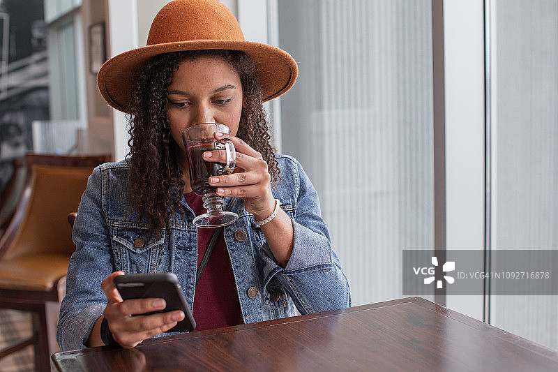 在机场的咖啡馆里，时髦的年轻女子一边喝着咖啡，一边看着智能手机。图片素材