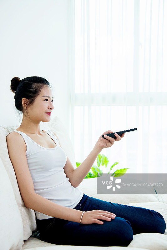 年轻的亚洲女人用遥控器在沙发上看电视图片素材