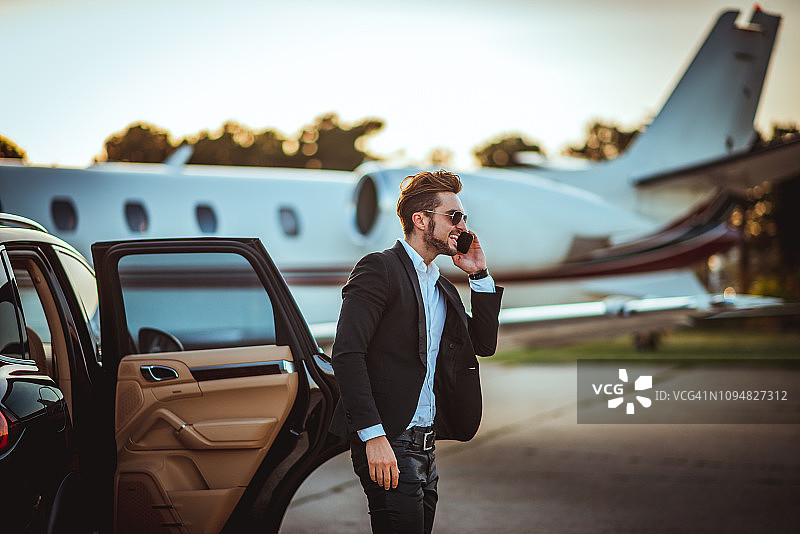 年轻的富商从停在停机坪上一架私人飞机旁边的豪华轿车里走出来，一边打电话图片素材
