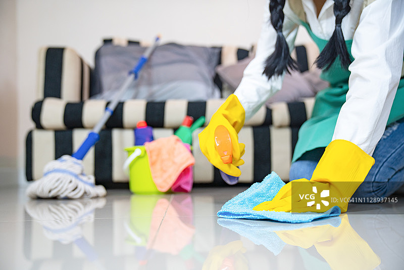 丈夫的家务和清洁概念，快乐的年轻女人戴着蓝色橡胶手套，用喷雾和抹布擦拭灰尘，同时打扫家里的地板图片素材