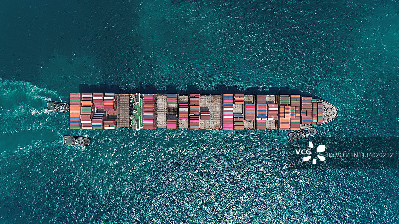 鸟瞰图大型集装箱船由三艘拖船拖着在港口卸货集装箱到仓库物流，进出口，海运或运输。图片素材