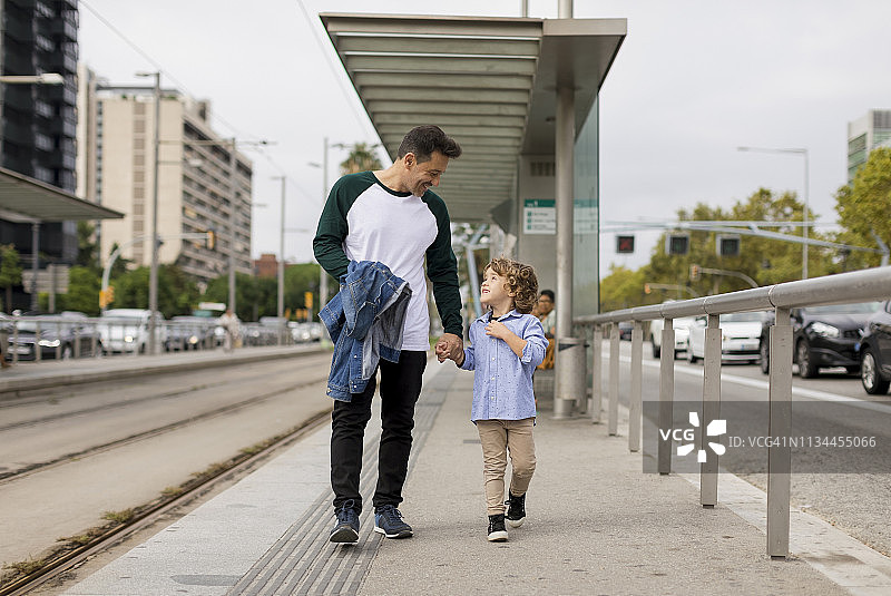 微笑的父亲和儿子手牵手走在城市的电车站图片素材