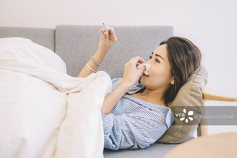 一名亚洲华裔女性躺在沙发上，穿着睡衣和毯子看着温度计图片素材