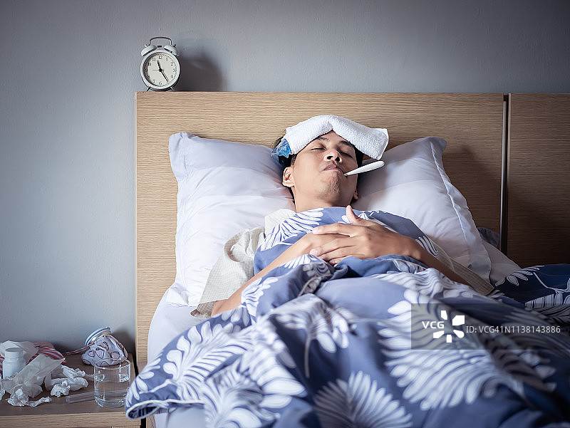 亚洲男子感冒高烧，睡在舒适的床上使用冰袋受伤减少和使用温度计测量温度。生病在家图片素材