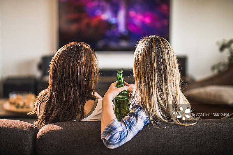 后视图两个女人放松在沙发上在家看电视。图片素材