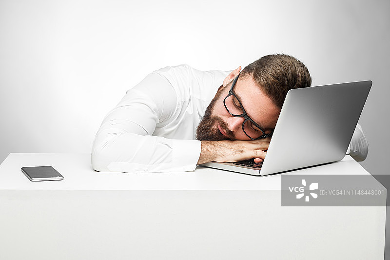 在工作站做个好梦。一个疲倦的自由职业者穿着白衬衫坐在办公室里，在笔记本电脑旁边打盹。图片素材