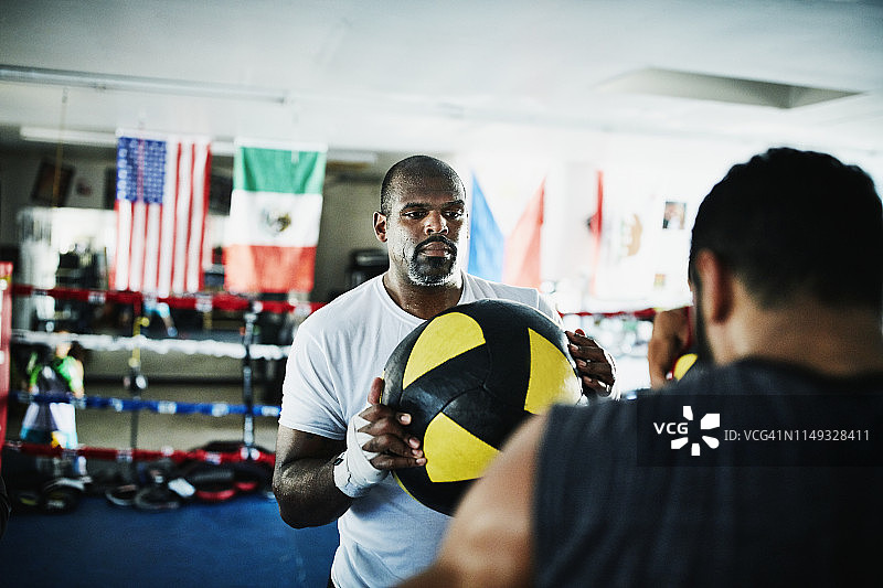 出汗的男性拳击手拿着实心球为伙伴打击训练期间在拳击馆的环图片素材