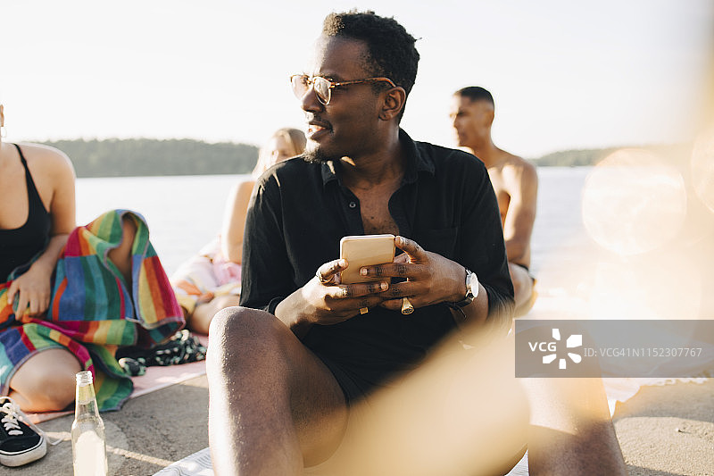 在夏天和朋友坐在码头上玩智能手机时微笑的男人图片素材