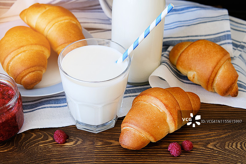 牛角面包装在盘子里，覆盆子果酱，牛奶装在瓶子里，牛奶装在玻璃杯里，用一根吸管盖在亚麻毛巾上，放在木桌上。健康饮食的概念。早餐或午餐食用有机农产品。图片素材