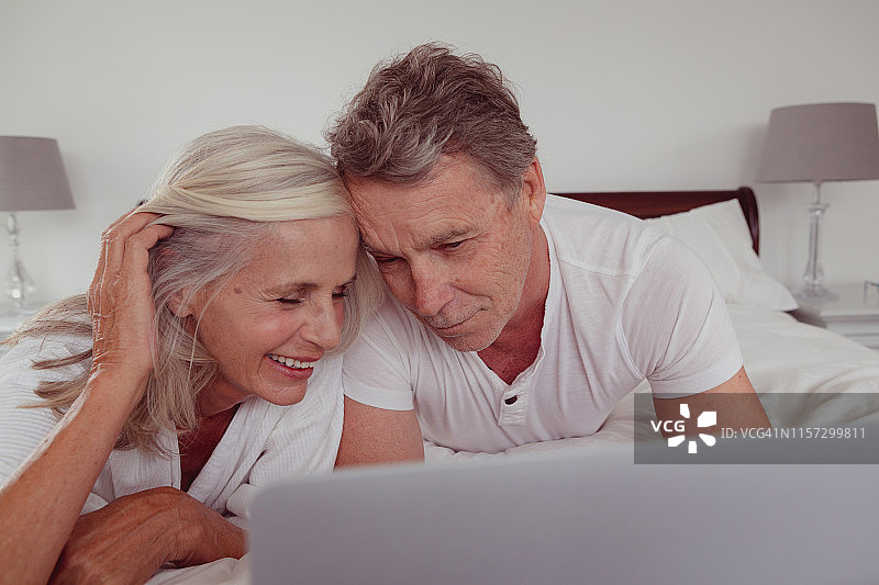 活跃的老年夫妇在舒适的家中躺在卧室的床上使用笔记本电脑图片素材
