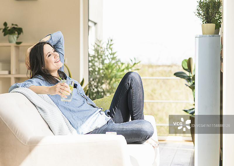一个快乐的女人，拿着一杯水坐在家里的沙发上图片素材