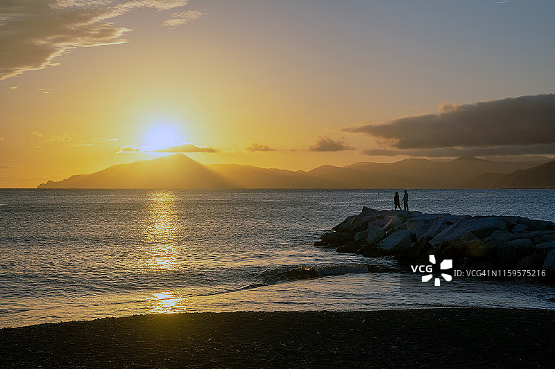 意大利，利古里亚里维埃拉，童话湾的背光海景，海滩上的岩石上有两个男人的剪影，日落时海面上地平线上的海岸线图片素材