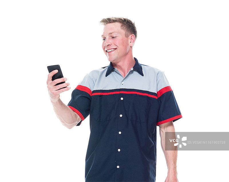 年轻英俊的男性机械师站在白色的背景上-在智能手机上发短信图片素材