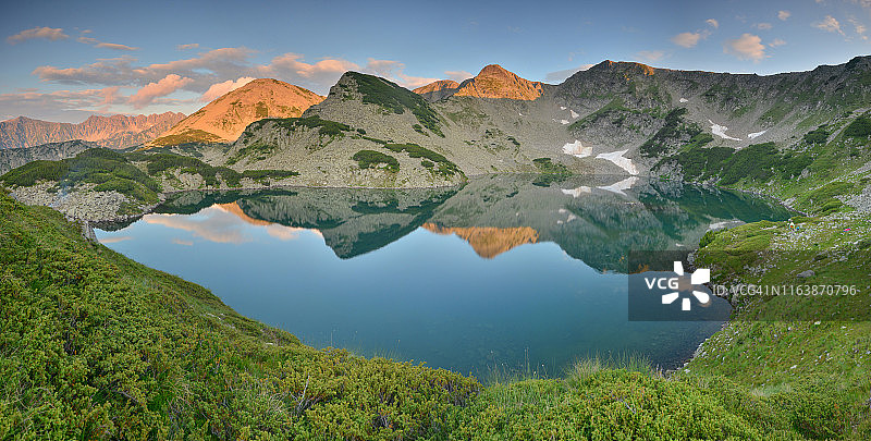 美丽的山湖与平静的水被绿色的草地和山峰上的朝霞包围的全景图片素材