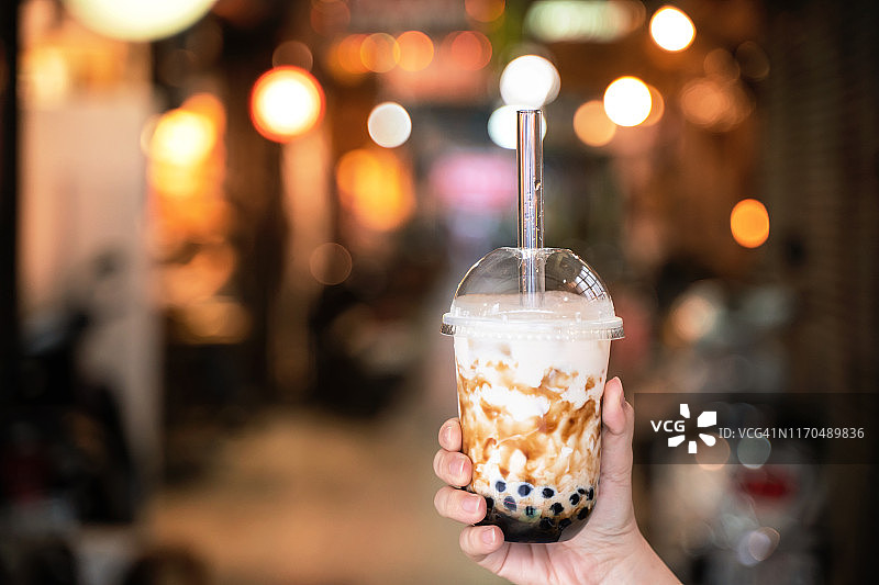 台湾夜市上，穿着牛仔夹克的小女孩正用玻璃吸管喝着红糖味的木薯珍珠奶茶。图片素材