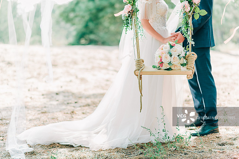 以新娘和新郎的手为背景，摆上装饰着鲜花和美丽的玫瑰花束的秋千。图片素材