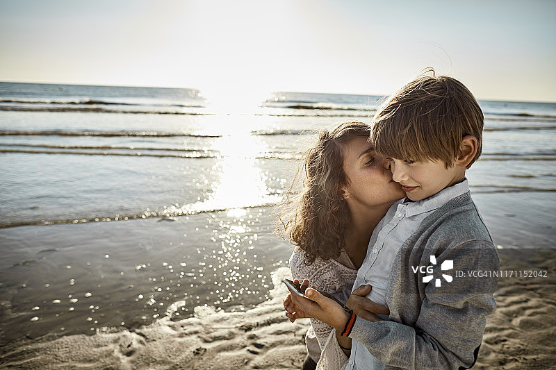 妈妈在沙滩上和拿着手机的男孩接吻图片素材
