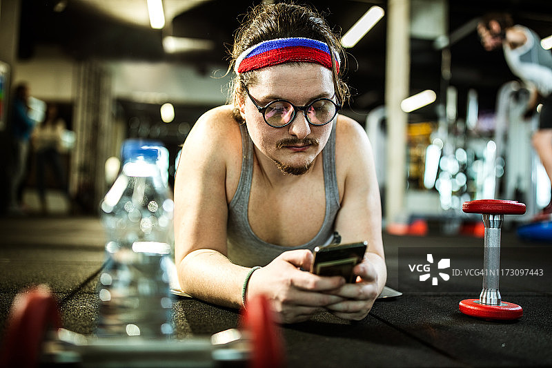 书呆子男运动员在健身房休息时用手机。图片素材