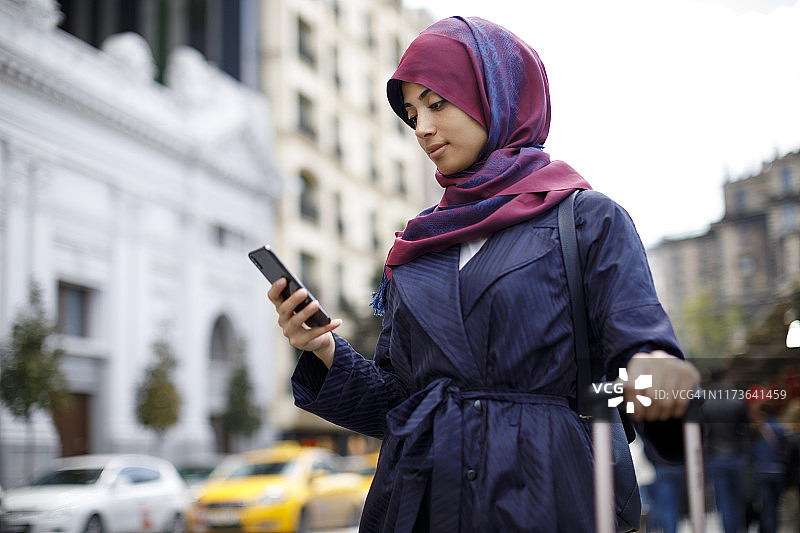 微笑的穆斯林妇女使用智能手机和等待出租车图片素材