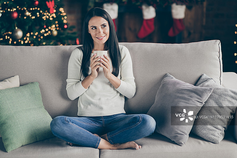 积极开朗的女孩的全身照片享受圣诞节庆祝假期思考计划思考fest拿杯热饮料坐沙发腿折叠享受新年传统在房子里与noel装饰图片素材